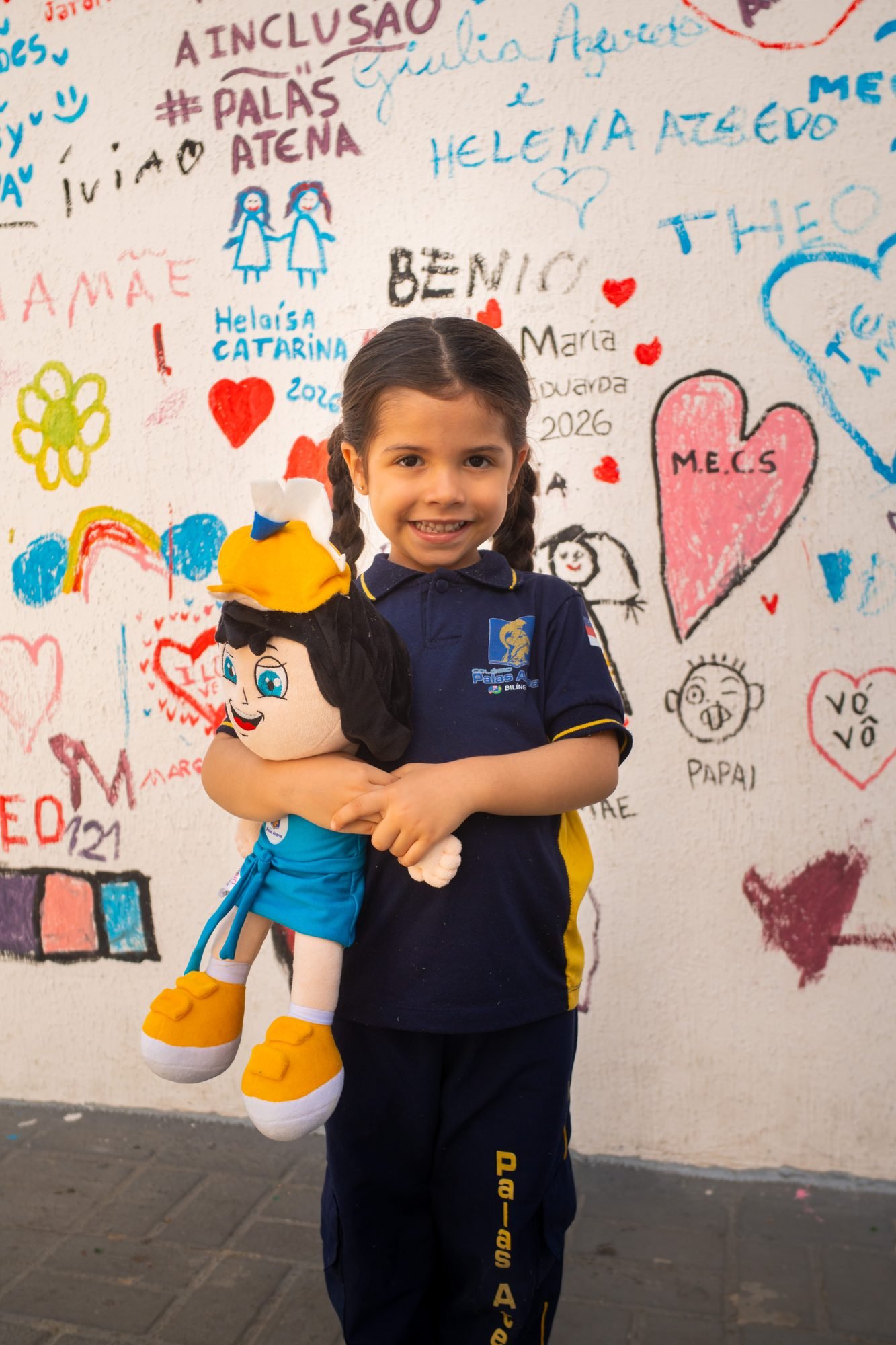 Aluna da Educação Infantil abraçada a uma boneca, diante do muro que os alunos pintaram